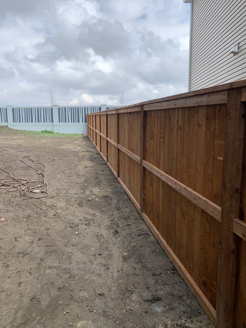 New wooden fence installation in progress alongside existing metal fence with construction site visi