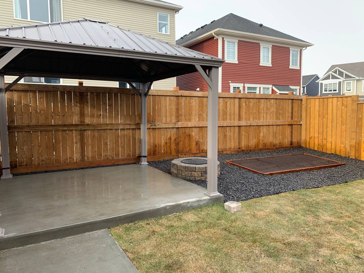 Completed backyard hardscape with concrete patio, pergola, wooden fence, fire pit, and gravel landsc
