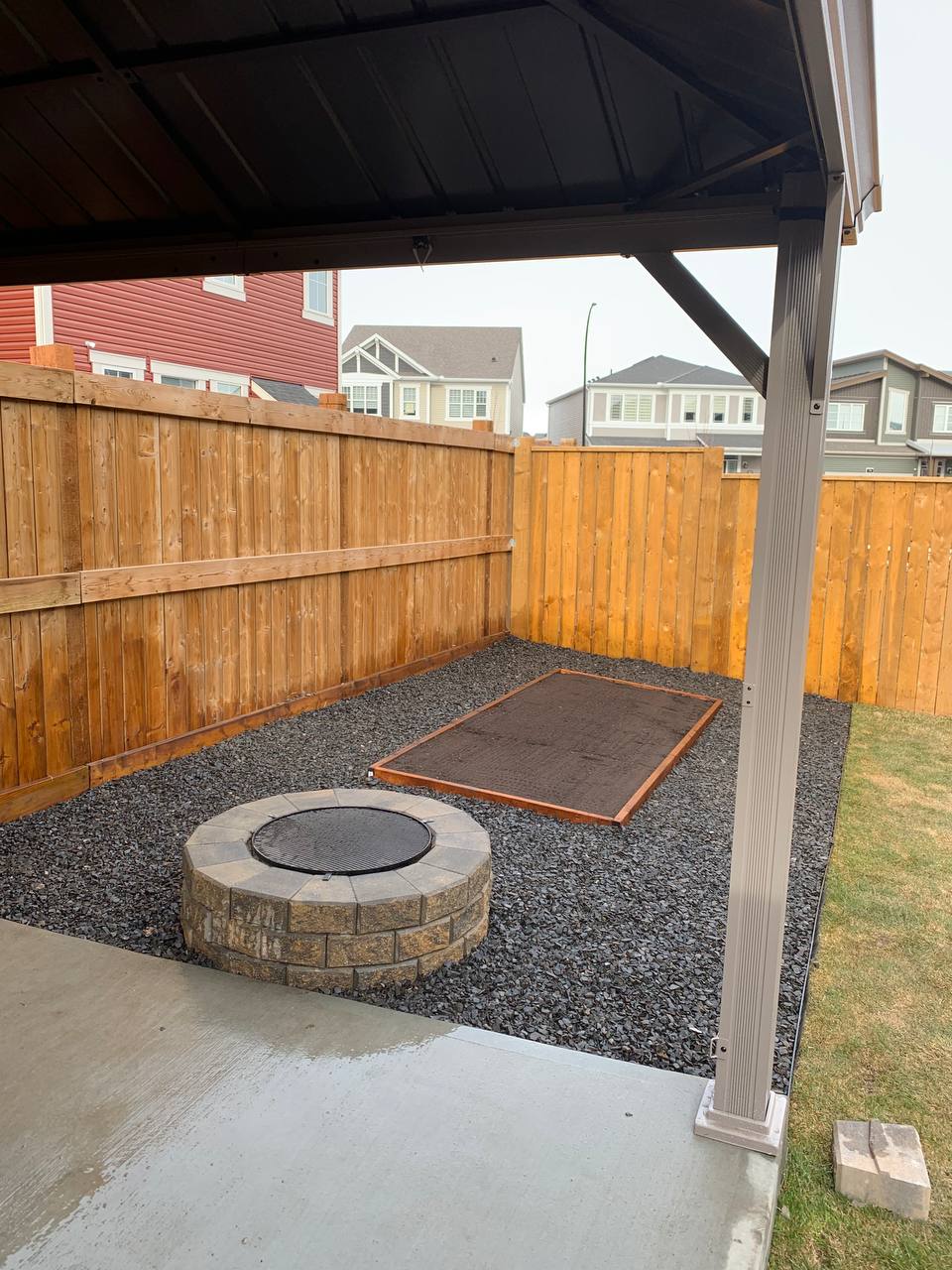 Completed backyard patio with gravel base, fire pit, cornhole board, and wooden fence under covered 